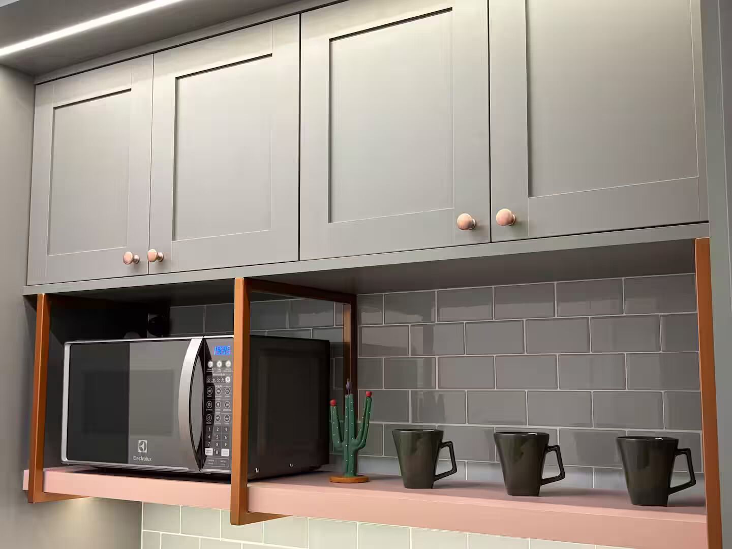 A kitchen with grey cabinets, a microwave, and three black mugs on a pink shelf.