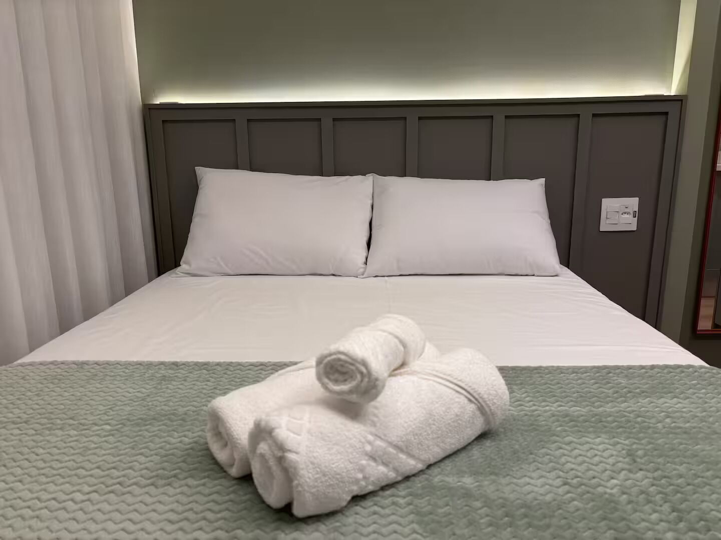 A neatly made bed with white linens and a rolled towel, a grey headboard, and a green textured bedspread.