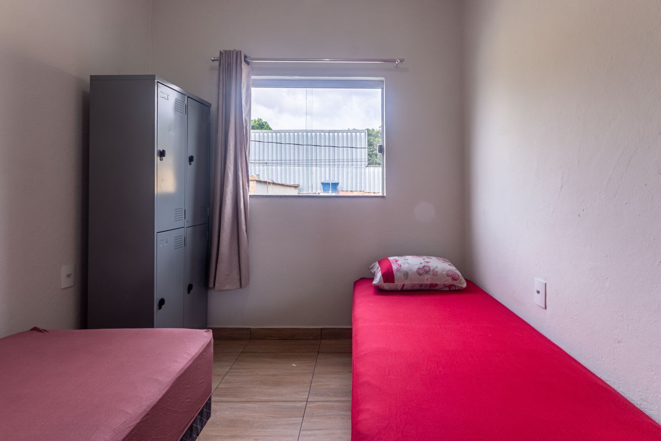 Ein kleines Zimmer mit einem Bett, einem Kleiderschrank und einem Fenster mit Blick auf einen Zaun und Bäume.