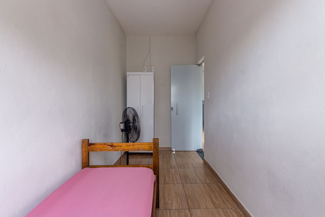 A narrow hallway with a bed on the left, a fan, and a door leading to a room.
