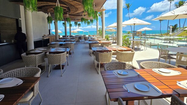 A covered outdoor dining area with wicker furniture, hanging greenery, and a view of the pool and ocean. - Cais Eco Residencia by Manta e BHL (Ipojuca)