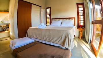 Quarto com cama, um banquinho, uma mesinha com um vaso e uma janela com cortinas.