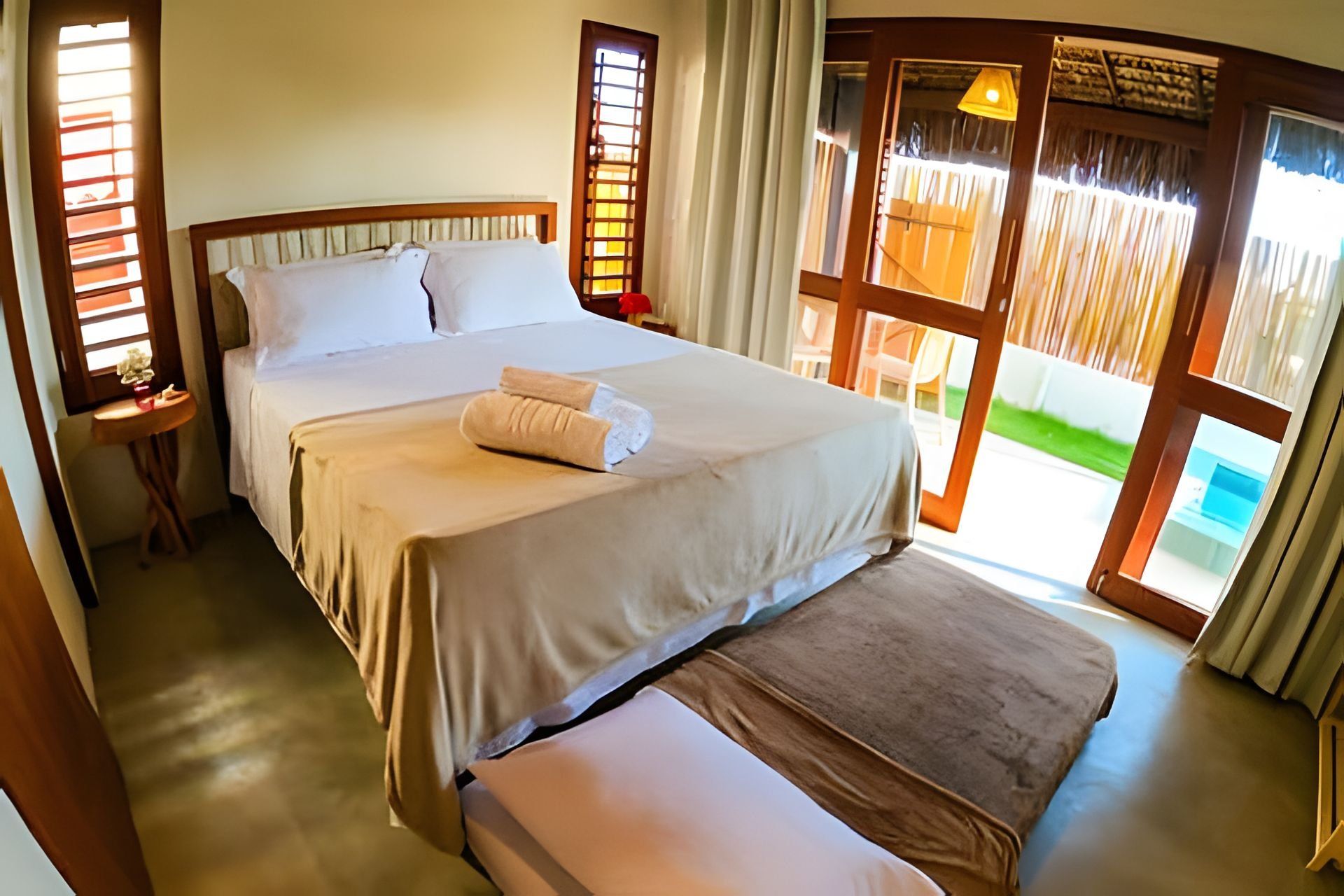 A double bed with a pillow and a rolled towel, a bedside table, and a window with blinds.