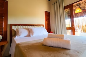 Una cama bien tendida con almohadas y una toalla doblada, un cabecero de madera, una ventana con cortinas y una lámpara colgante.
