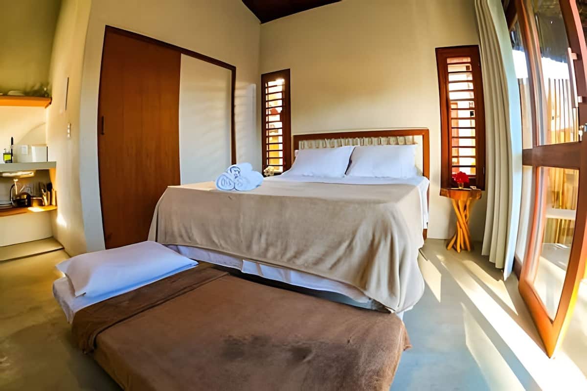 A bedroom with a bed, a bench, a small table with a vase, and a window with curtains.