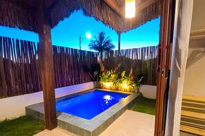 Una piscina con muelle de madera, rodeada por una valla y plantas, bajo un techo de paja.