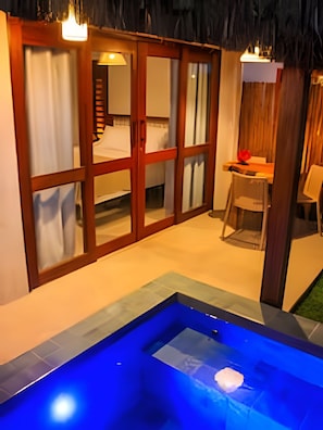 Una habitación con puerta de vidrio corredera que da a un balcón, una mesa pequeña con una silla y una zona de piscina iluminada en azul.
