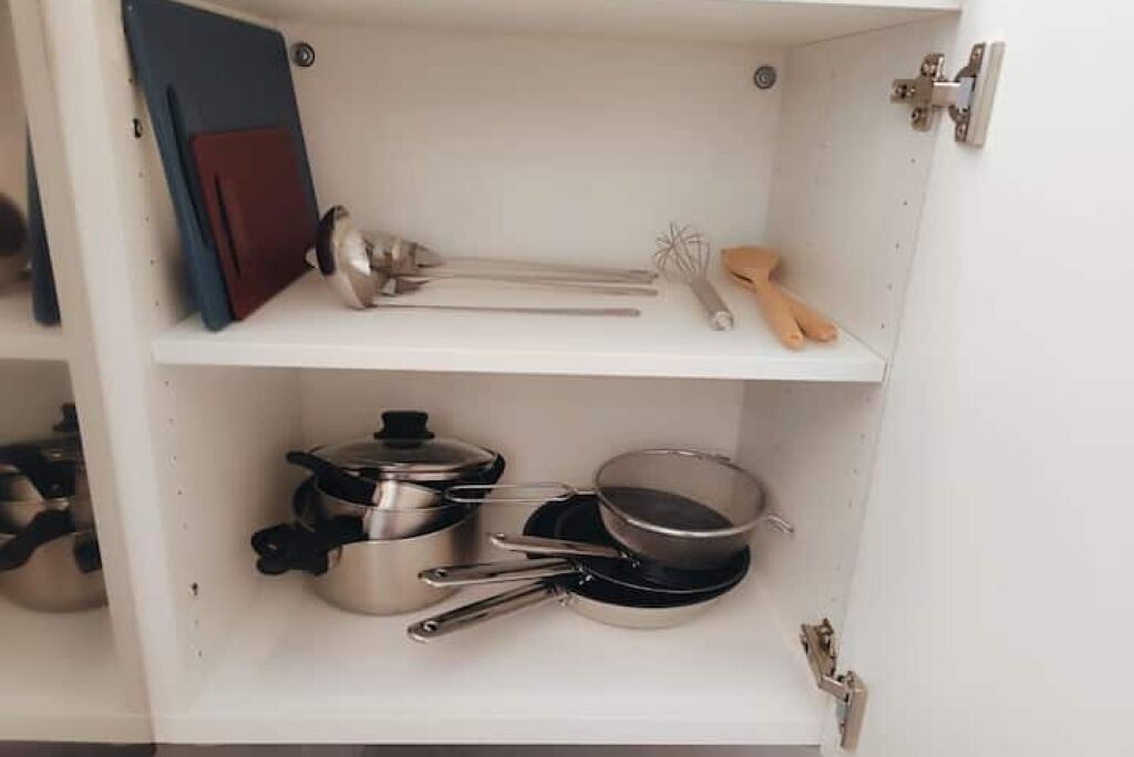 A kitchen cabinet with stainless steel cookware, a whisk, and a red folder.