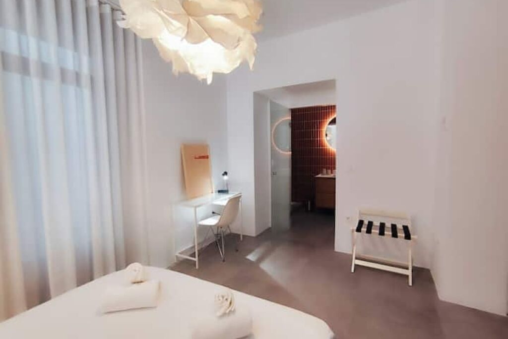 Una habitación de hotel moderna con una cama, un escritorio, una silla, un taburete pequeño y una lámpara de techo original.