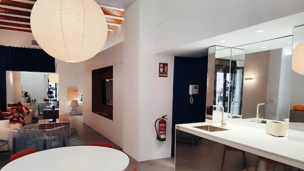 Un hall d’hôtel moderne avec une table de salle à manger ronde, des chaises rouges, un grand luminaire suspendu, un extincteur et un mur recouvert de miroirs.