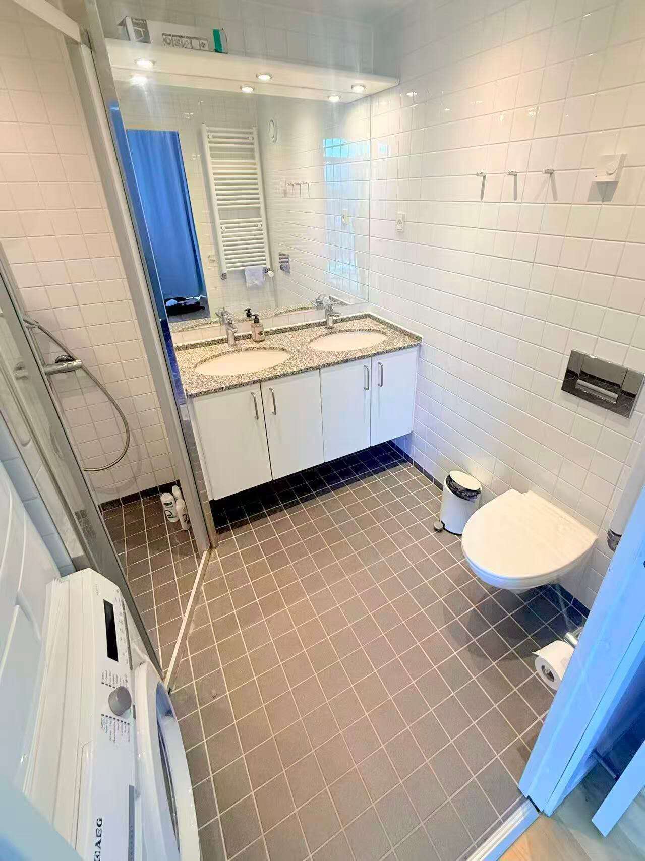 Ein Badezimmer mit weißen Fliesen, einem Doppelwaschbecken mit geflecktem Ablagetisch, einer weißen Toilette und einer Duschkabine mit Glastür.