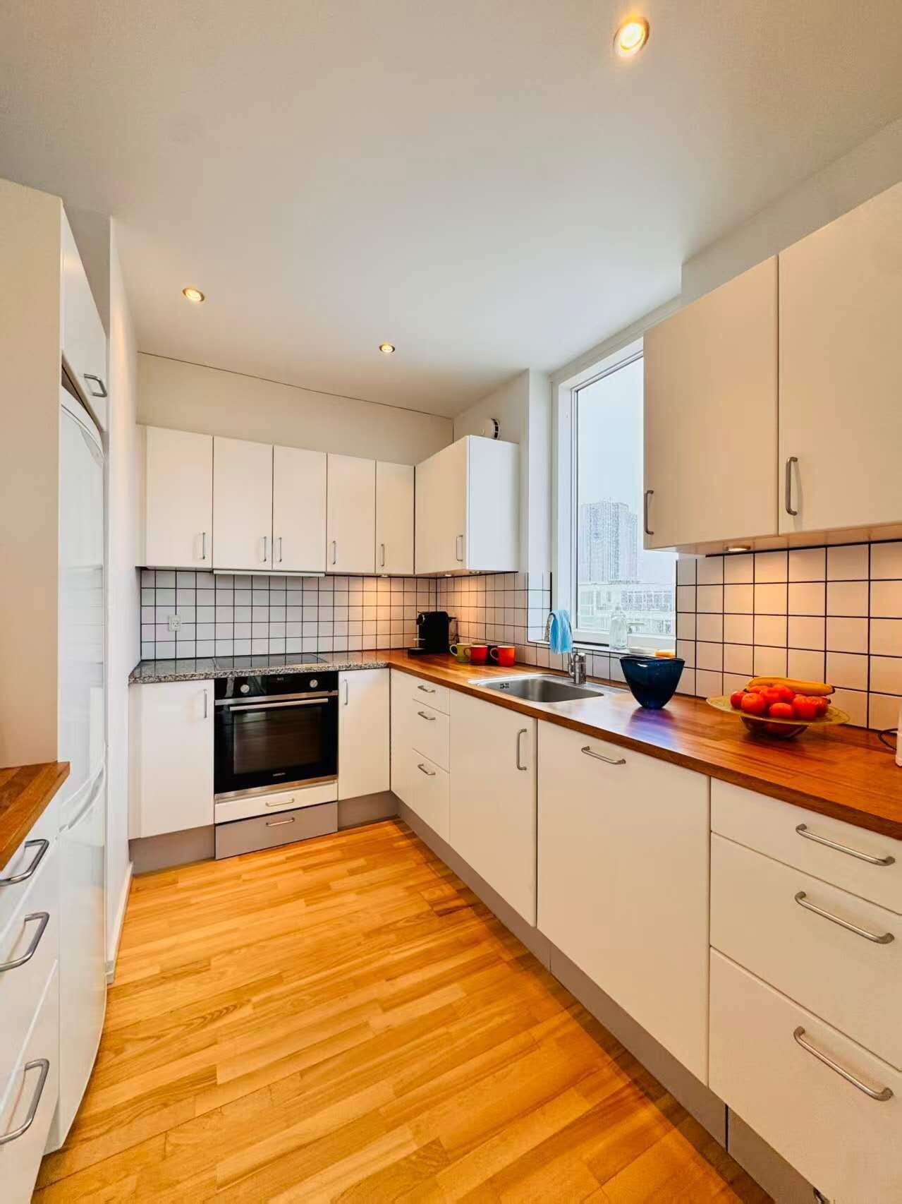 A modern kitchen with white cabinets, a wooden countertop, a built-in oven, and a window providing a city view.