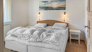 A double bed with white bedding, a wicker headboard, two bedside tables with lamps, a white radiator, and a painting of a seascape above the bed. - Beautiful home in Blokhus with WiFi (Blokhus)