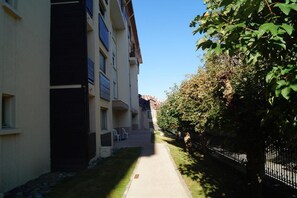 Ein schmaler Weg zwischen Gebäuden, ein baumbestandener Zaun und ein klarer Himmel.