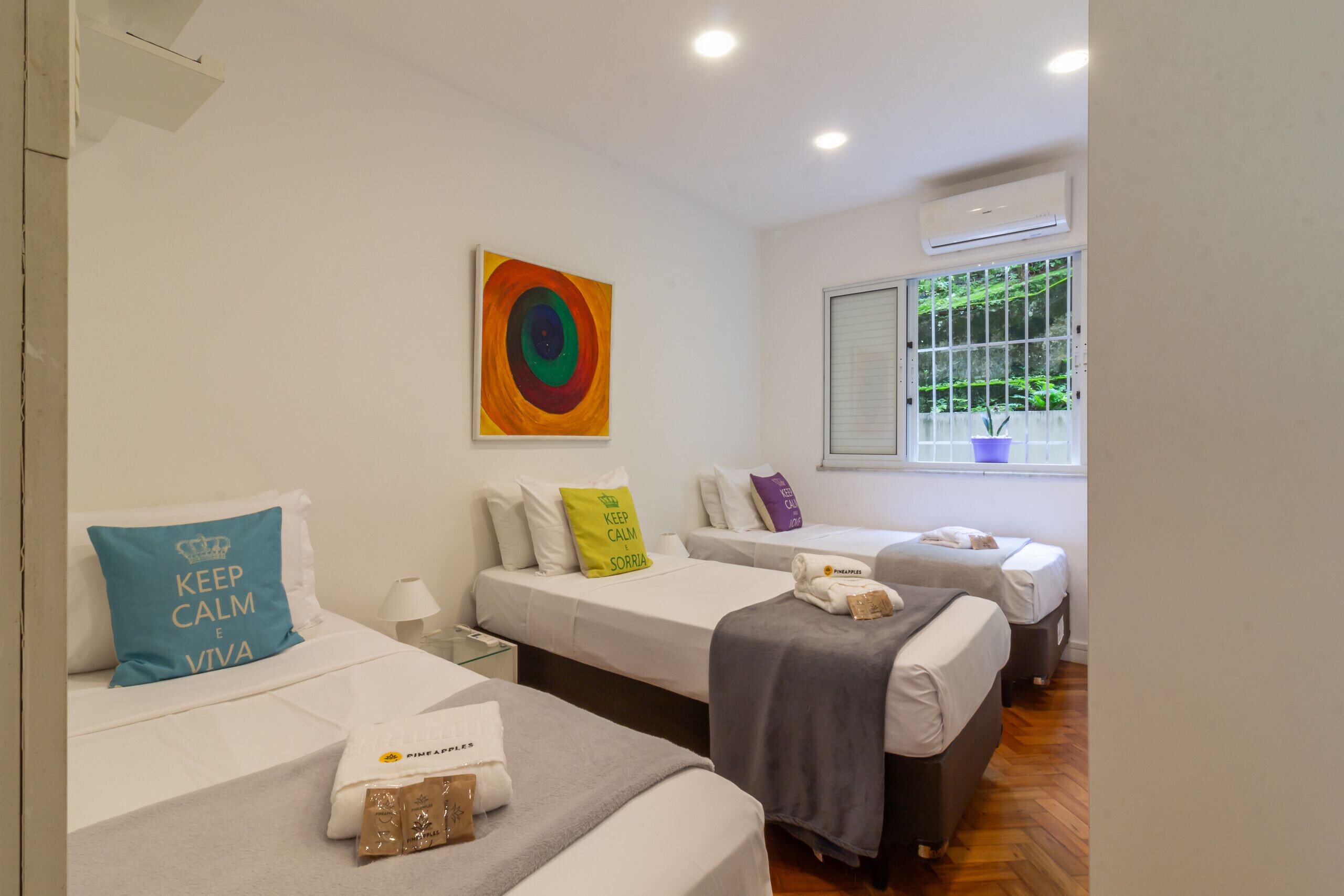 A hotel room with two beds, a window with a view of greenery, an air conditioner, and a colorful painting on the wall.