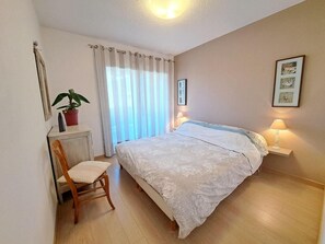 Ein Schlafzimmer mit einem Bett, einem Sessel, einer Zimmerpflanze, einem Fenster mit Vorhängen und einem Wandbild.
