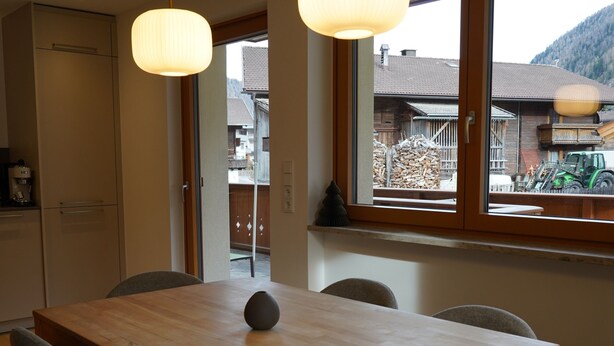 Un comedor con una mesa de madera, cuatro sillas grises, dos lámparas colgantes y una ventana con vistas a una montaña y un tractor.