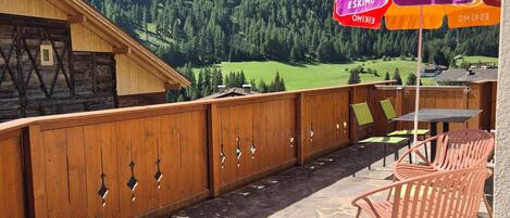A wooden deck with outdoor seating, a view of a mountainous landscape, and a wooden building in the background.