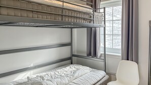 A bunk bed with a white mattress and a single bed below, a white chair, and a window with curtains.