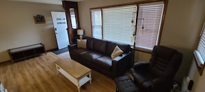 Una sala de estar con un sofá de cuero marrón, una mesa de centro de madera, un sillón negro y una ventana con persianas.