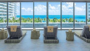 Una stanza con una grande finestra vista spiaggia e edifici di un hotel, con poltrone relax e un piccolo tavolo.