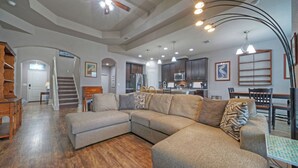 A spacious living room with a sectional sofa, a dining area with a table and chairs, a kitchen with dark cabinets, and a staircase leading to another level. - Rhumba by Avantstay Game Room + Bakyard Oasis + Outdoor Dining Near Beaches and Downtown (Corpus Christi)