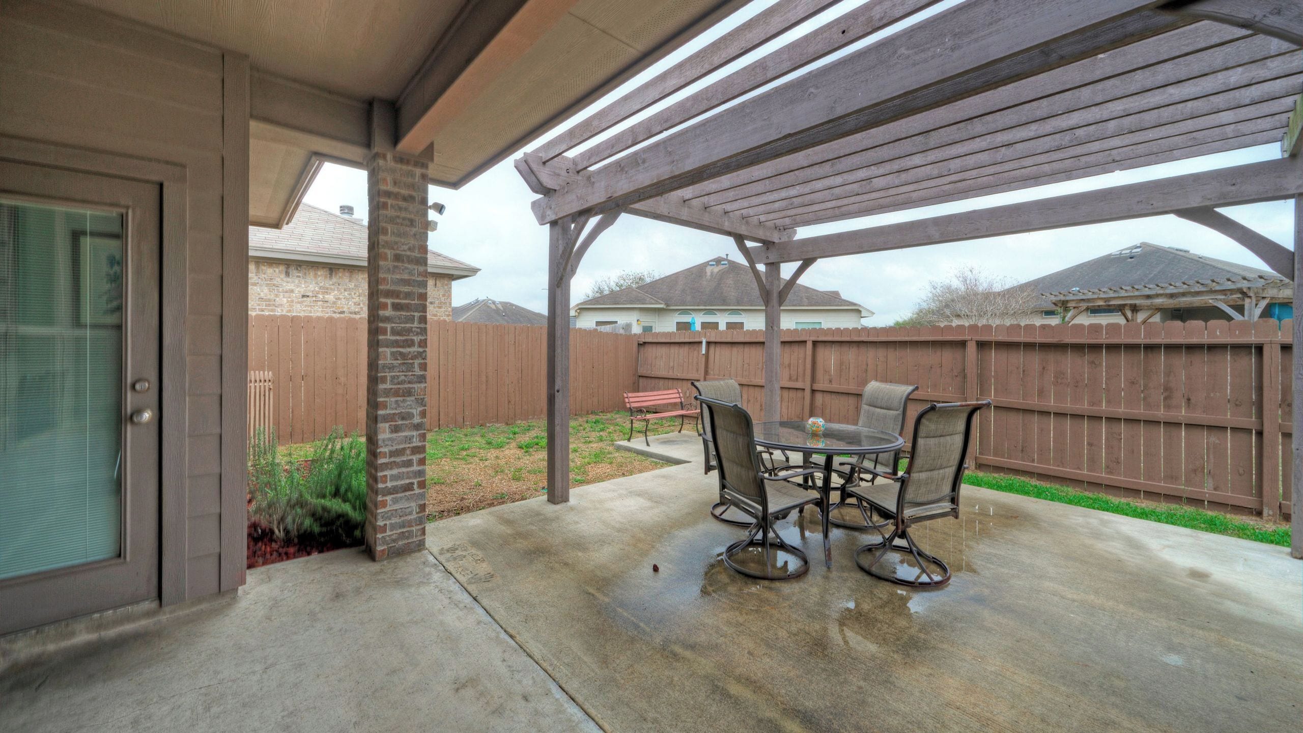 A covered patio with a table and chairs, a wooden fence, and a house in the background.