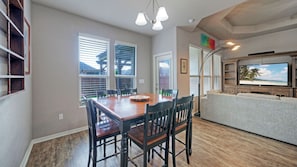 A dining area with a wooden table and chairs, a television in the living room, and large windows with blinds. - Rhumba by Avantstay Game Room + Bakyard Oasis + Outdoor Dining Near Beaches and Downtown (Corpus Christi)