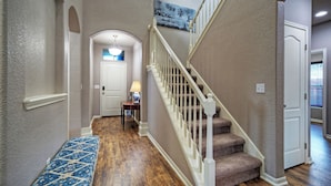 A hallway with a wooden floor, a blue patterned rug, a white door, a staircase with white railings, and a light fixture. - Rhumba by Avantstay Game Room + Bakyard Oasis + Outdoor Dining Near Beaches and Downtown (Corpus Christi)