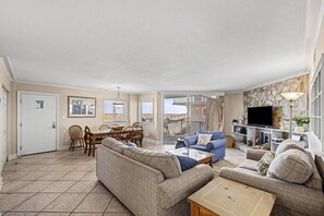 A spacious living area with a sofa, coffee table, and TV. There's a dining area with chairs and a table. The room has a balcony with a view. - Spacious Gulf Front Condo - SV 111 (Pensacola Beach)