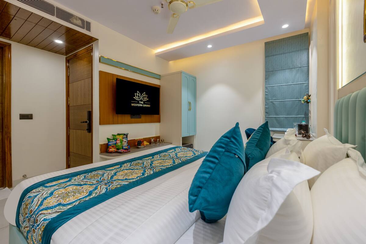 A hotel room with a bed, a TV mounted on the wall, a window with blinds, and a ceiling fan.