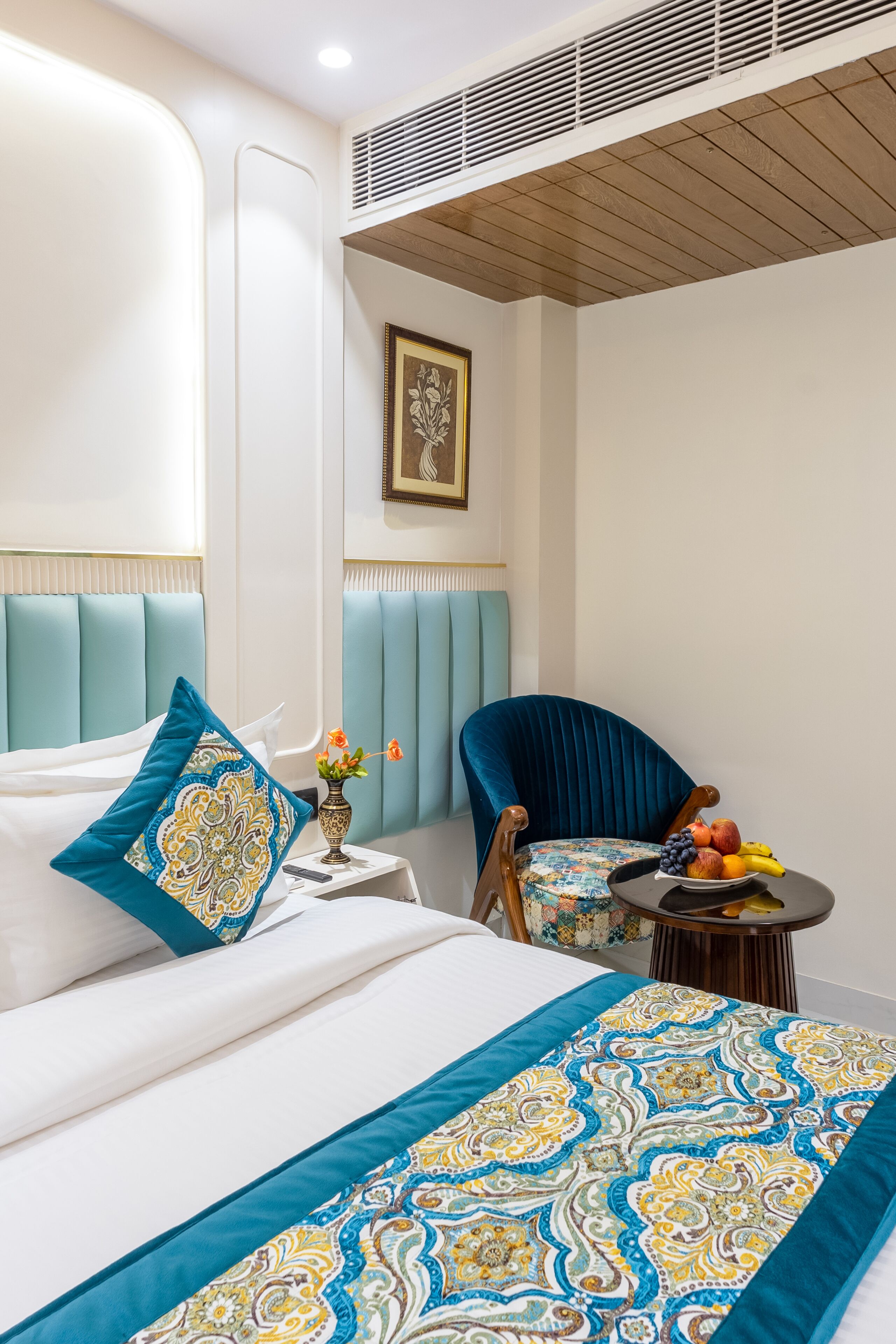 A hotel room with a bed, a chair, a small table with fruit, and a vase with flowers.