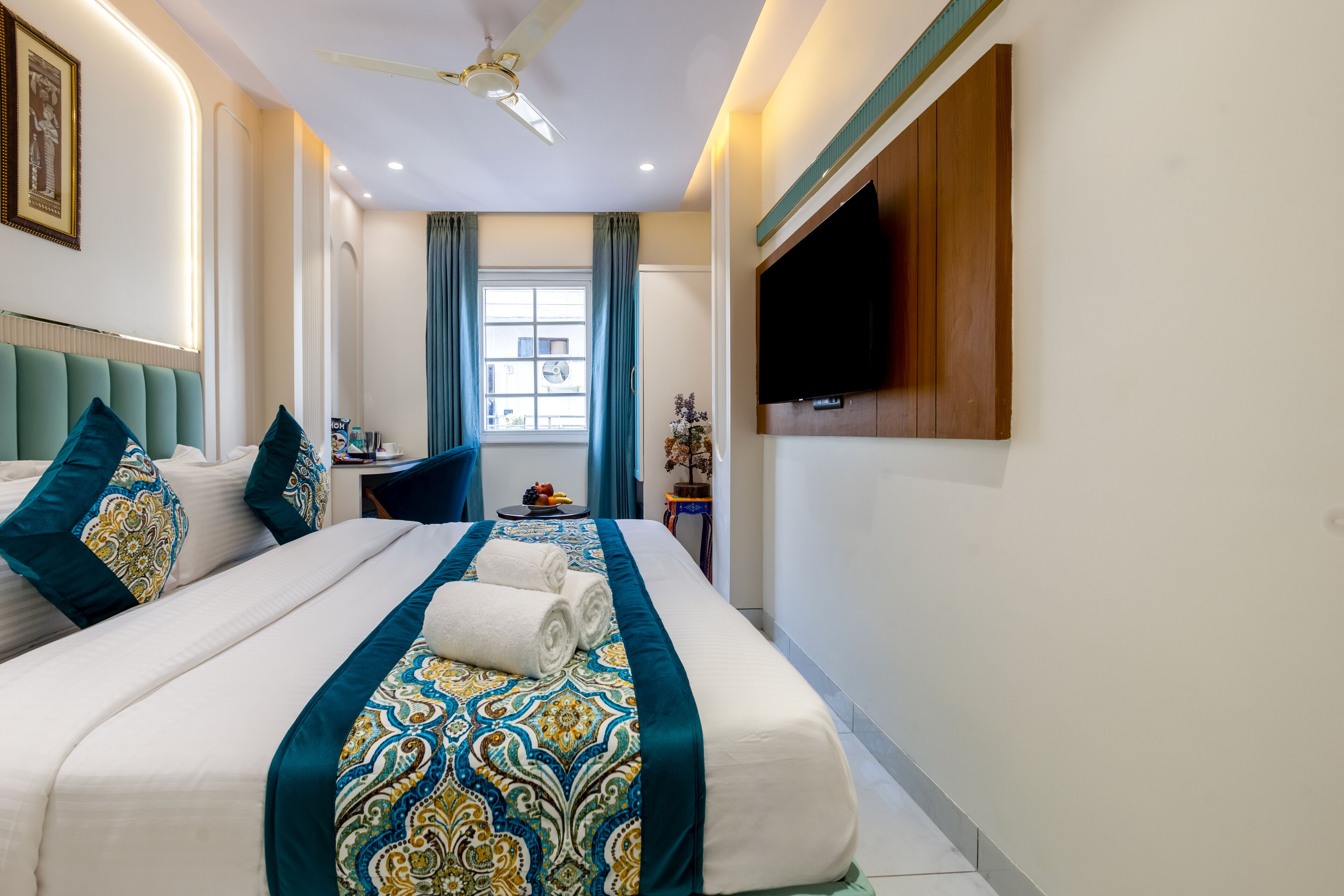 Habitación de hotel con una cama grande, ventilador de techo, televisor montado en la pared y una ventana con cortinas.