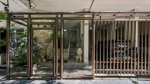Uma entrada protegida por grades, com uma porta de vidro e uma escadaria que leva ao interior, além de um pavimento ladrilhado.