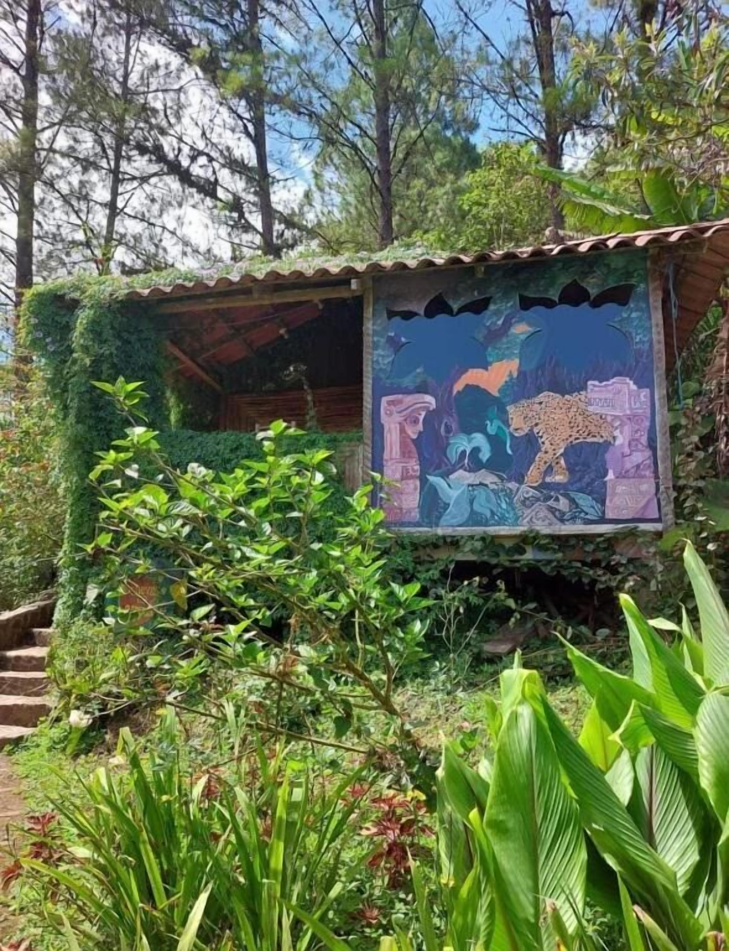 A small, rustic hut with a mural of a leopard and a bird, surrounded by lush greenery and trees.