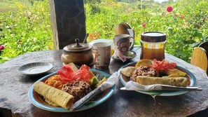 Una mesa con platos de comida, un cebrero, una taza y un termo, todo sobre una mesa con vista a un jardín.