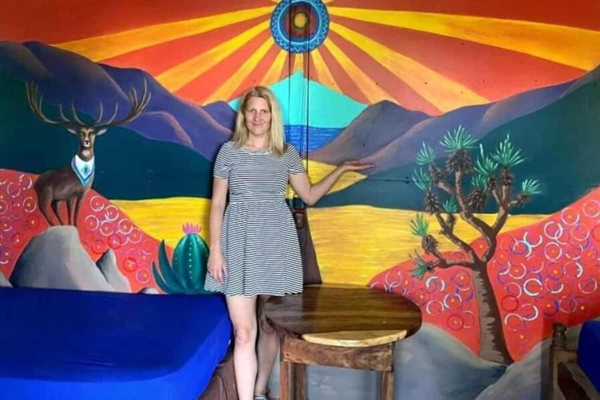 Une personne se tient devant une fresque murale colorée représentant une montagne, un arbre et un ornement suspendu.