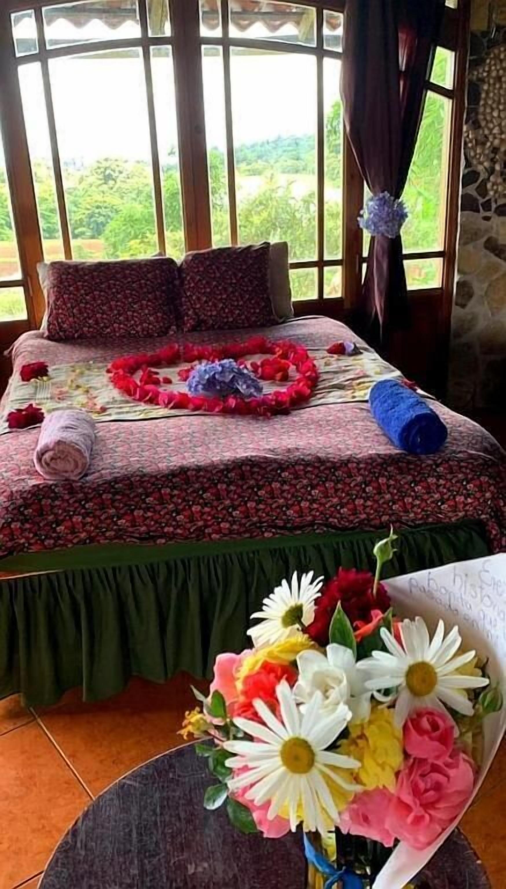A bed with a floral bedspread, a rolled towel, and a bouquet of flowers.