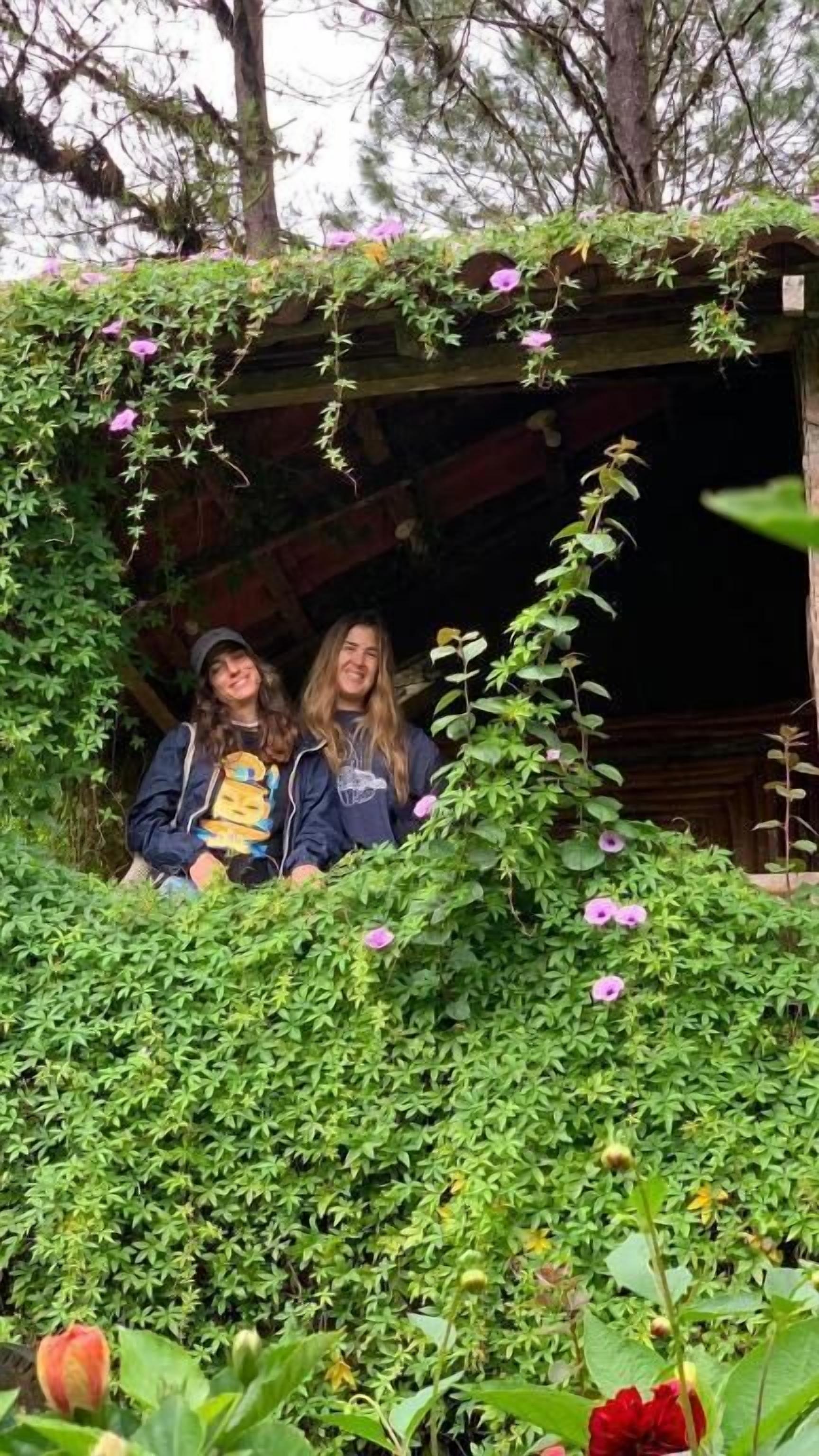 Two individuals in a garden area with dense foliage and a wooden structure.