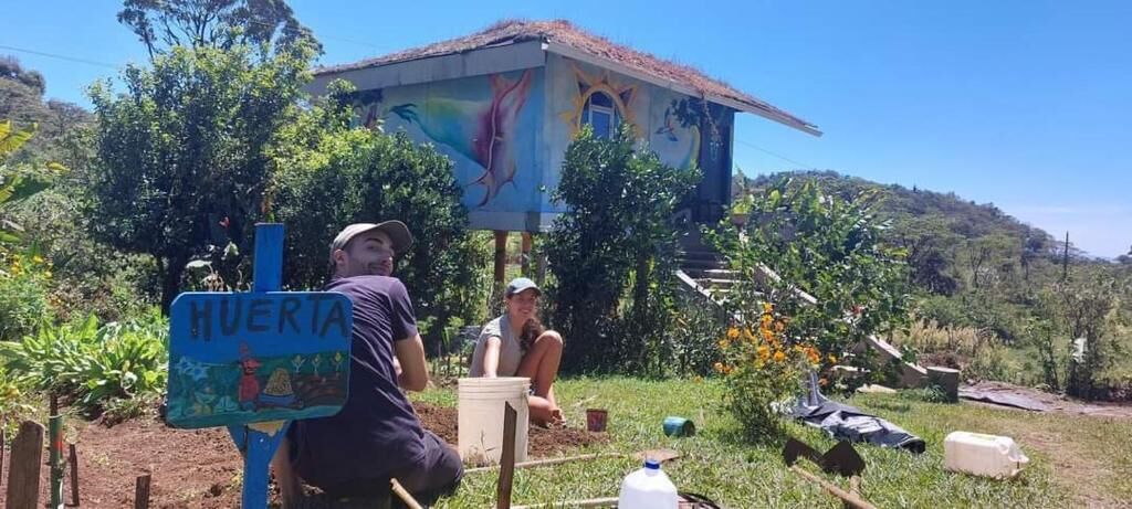 Edificio rústico con un mural, un letrero que dice 'HUERTA' y una persona sentada en una silla.
