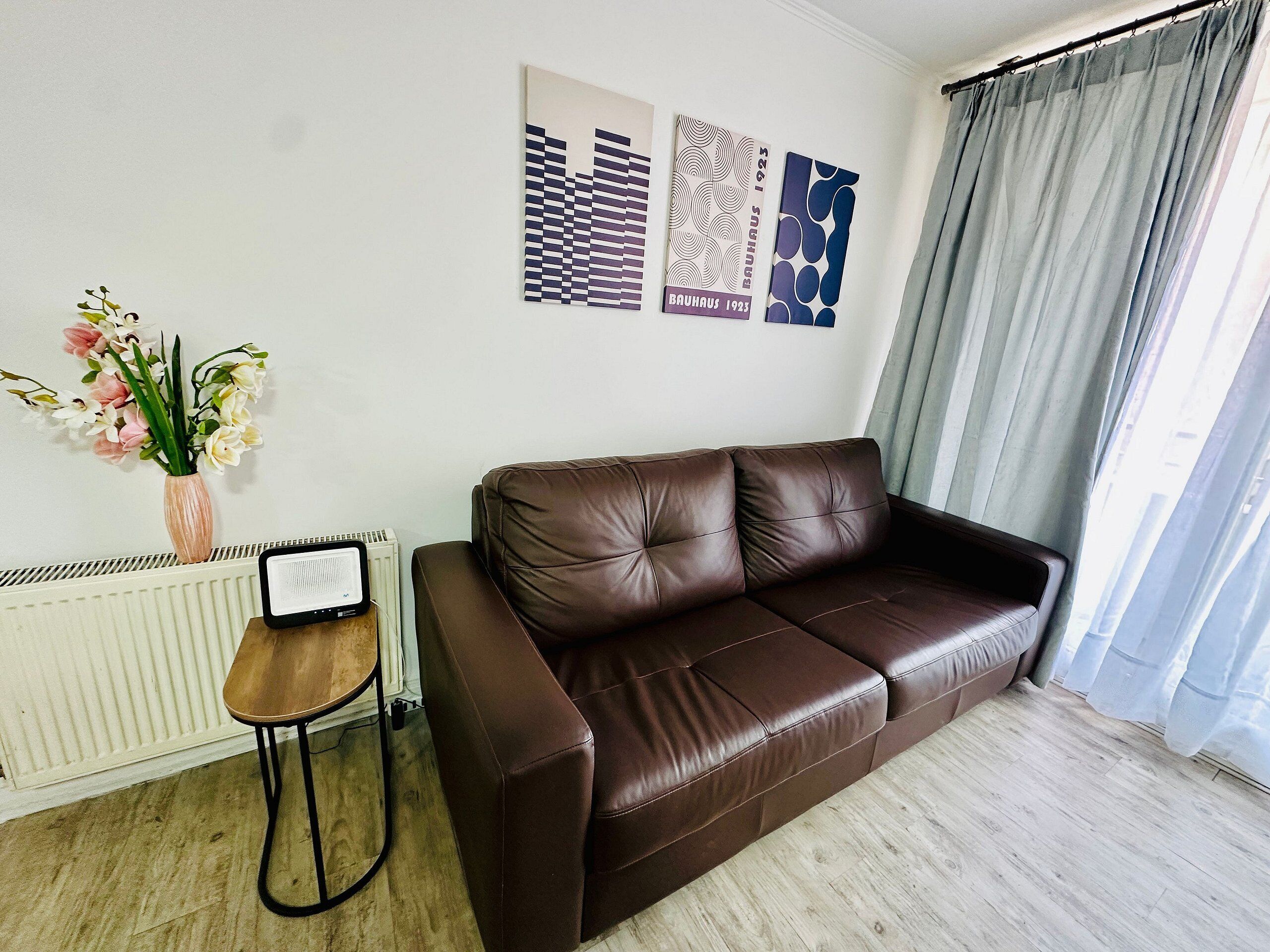 Een bruine leren bank, een radiator, een klein houten krukje en een vaas met bloemen.