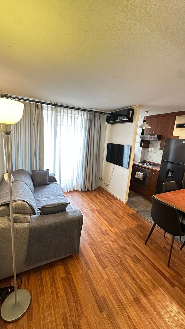 A living room with a sofa, a dining table, a TV mounted on the wall, and a kitchen area with a stove and refrigerator.