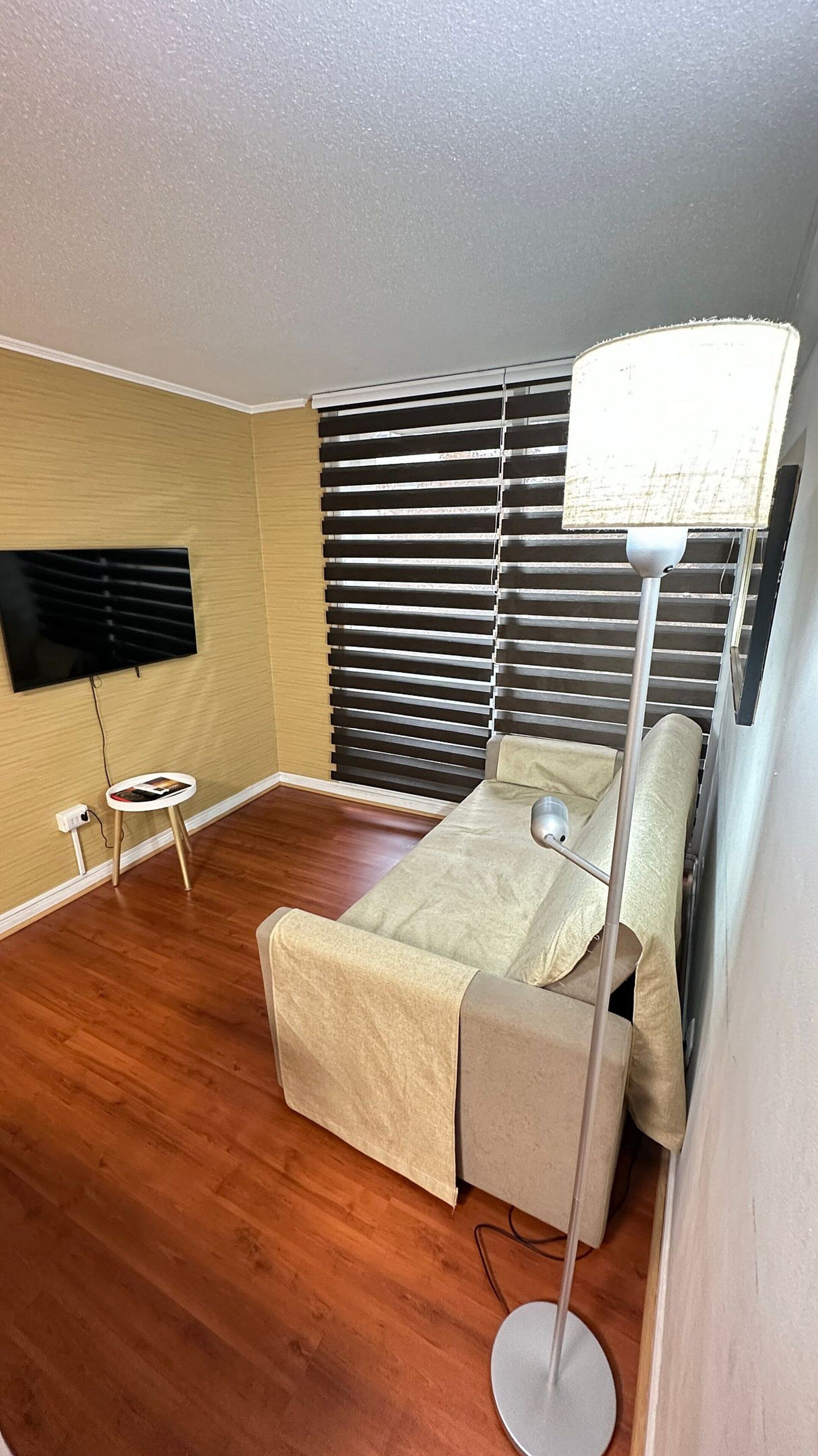 Habitación pequeña con cama, lámpara de pie, televisor montado en la pared y una mesita.
