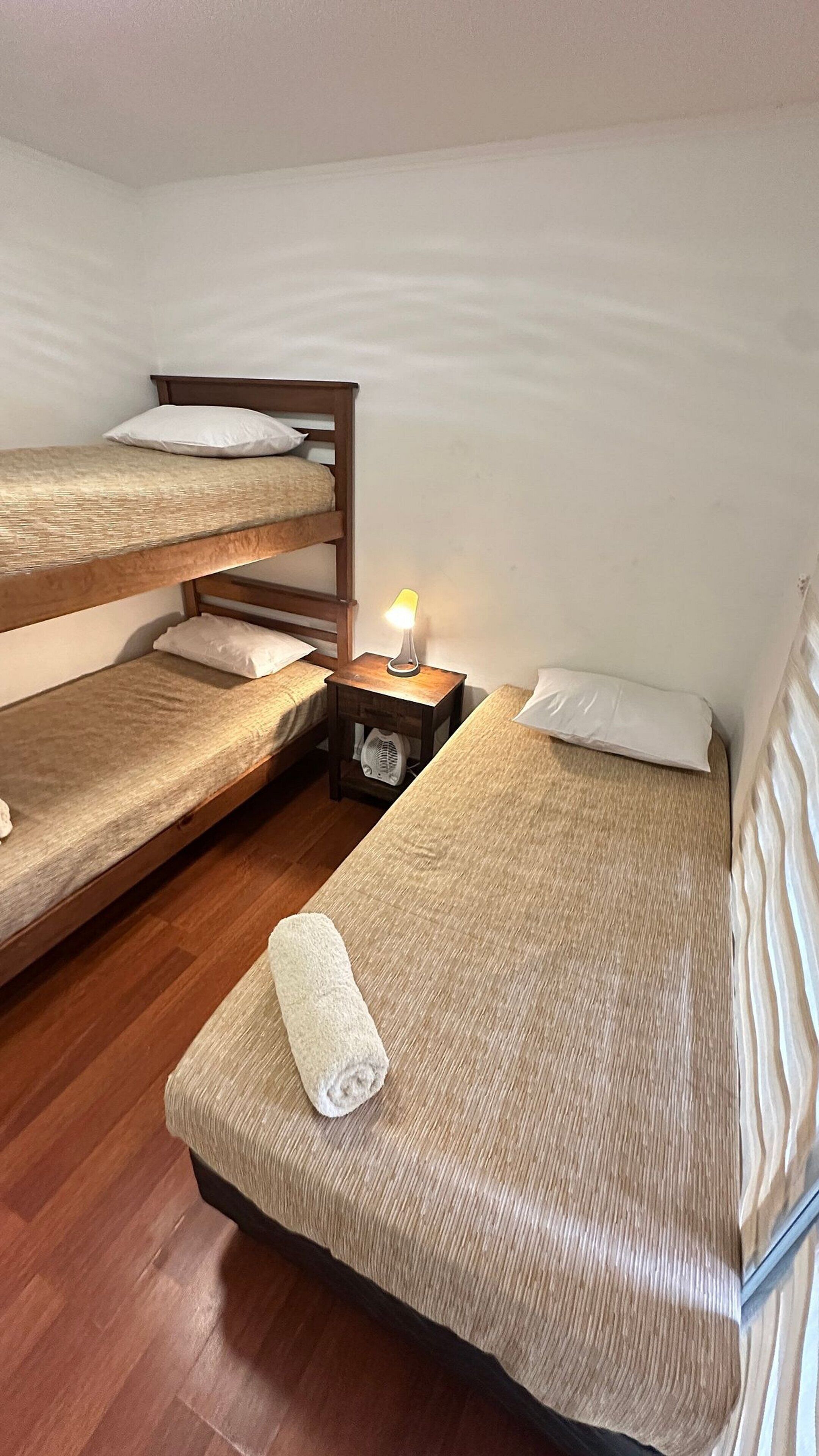 A room with three bunk beds, a bedside table with a lamp, and a rolled towel on the floor.