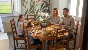 Une famille est assise autour d’une table ronde en bois, entourée de chaises. Un mur est décoré d’une fresque florale, et une fenêtre offre une vue sur un champ verdoyant.