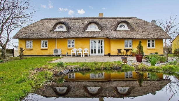Ein gelbes Haus mit einem Strohdach, vor der Fassade ein Teich, und ein klarer Himmel.