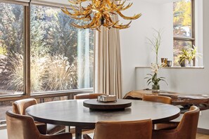 A round dining table with chairs, a chandelier, and a view of trees through large windows. - Charming 5-Bedroom Home w/ Private Spa and A/C in Beautiful West Vail (Vail)
