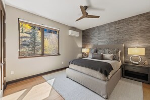 Un dormitorio con una cama, una mesita de noche, aire acondicionado de pared, ventilador de techo y un amplio ventanal con vistas a árboles.