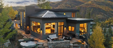 A modern two-story house with large windows, a stone patio, and a view of the surrounding hills.