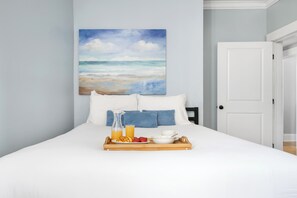 Ein ordentlich bezogenes Bett mit weißen Leinen, ein Tablett mit Frühstücksangeboten und ein Strandbild an der Wand.